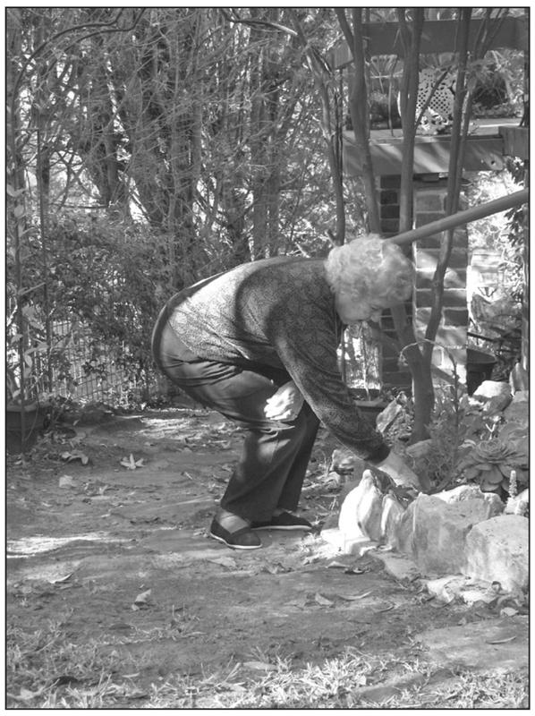 A black and white photograph of an elderly woman bending down with her knees to pull something from her garden.