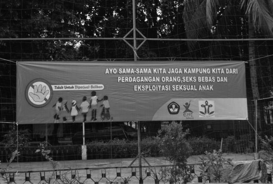 A black and white photograph of a banner hung along a wired fence. This banner states ‘Come on, together we protect our village from human trafficking, free-sex and child sexual exploitation’.