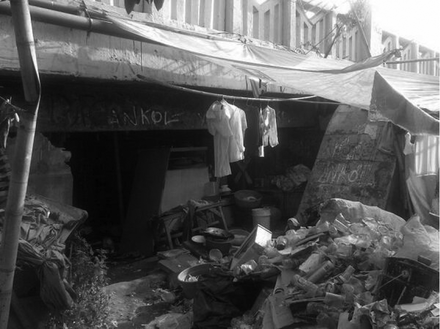 A black and white photograph of a slum area; piles of rubbish sit outside the entrance of a house. 