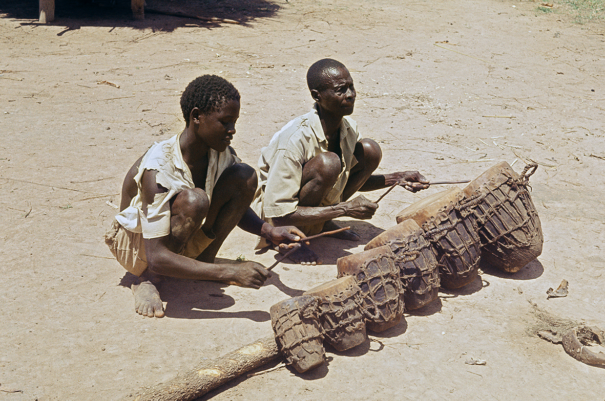 Figure 6.5. A middle-aged man and his son crouch on the floor, holding sticks which are used to drum on six different-sized drums that are tied to one another and to a long branch which lies on the dusty ground.