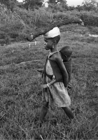 Figure 7.6. In a black and white photograph, a young girl of about 12 carries a toddler on her back while balancing a branch on her head.
