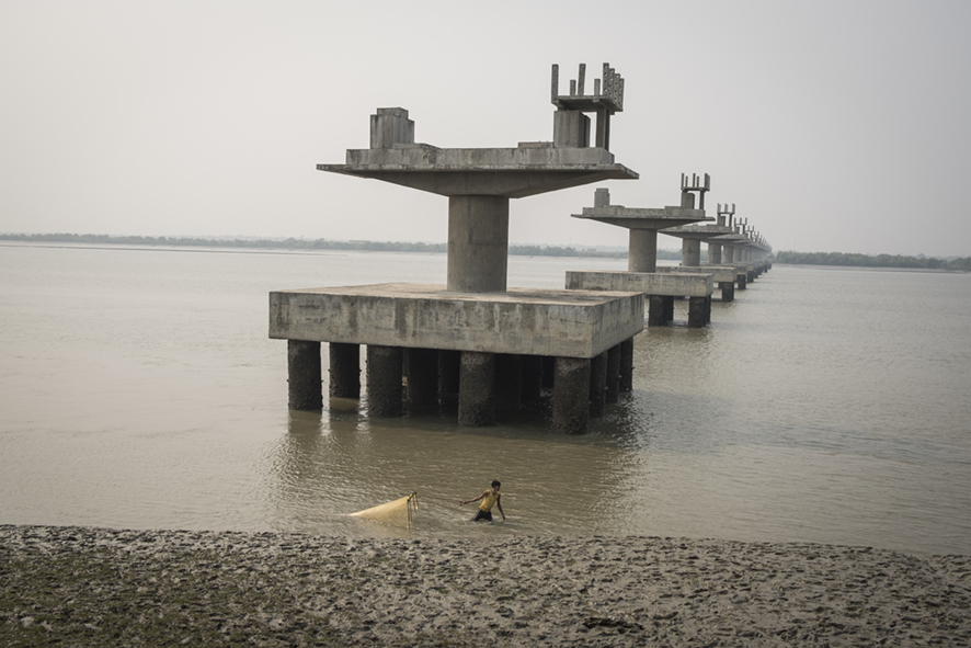 Man fishing with net in the Hooghly River, India with unfinished construction and exposed concrete pillars in the water behind him.