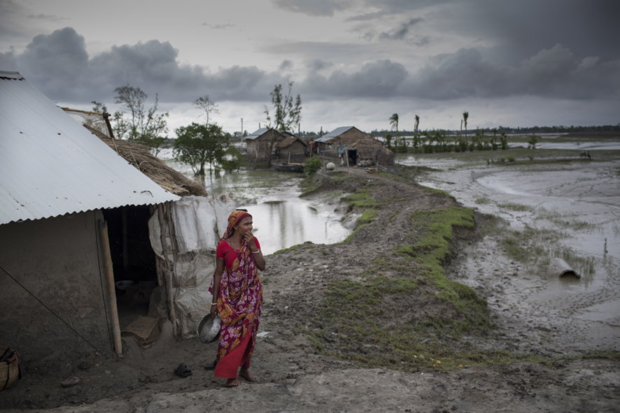 Bangladeshi woman in a red and purple sari outfit standing next to a river, surrounded by mud.