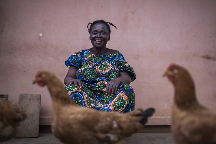 Central African Republic woman in vibrant patterned blue, orange and green dress, sitting down and smiling. Chickens walking by in the foreground. 