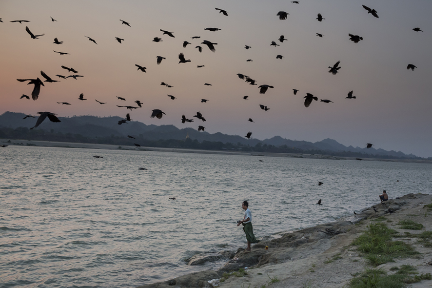 Birds flying over Irrawady River in Myanmar while a man is fishing by the shoreline in the distance.
