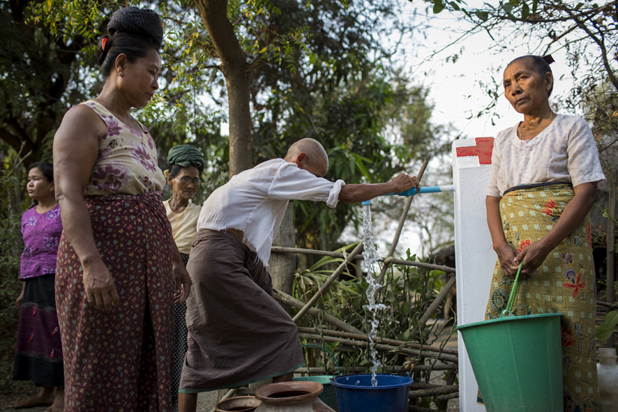 Elderly man filling up bucket at a communcal tap close to his home in central Myanmar. Two woman are in the foreground appearing to be waiting for their turn. 
