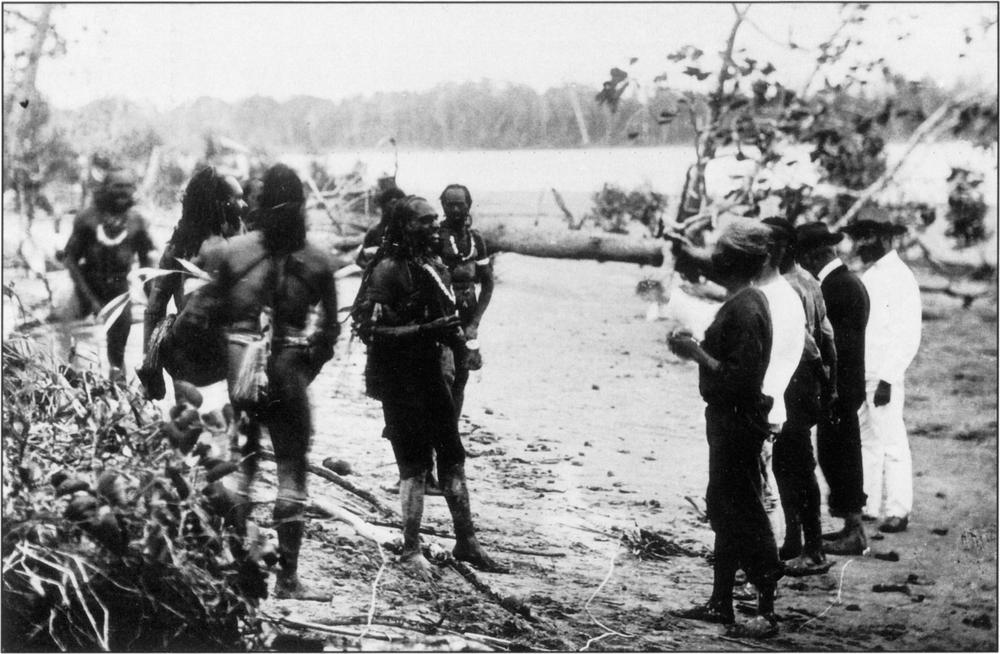Black-and-white photograph of Marind-anim men interacting with Dutchmen in an outdoor area of natural landscape. All of them appear to be bare-footed and standing on dirt ground with patches of sediment in sight, as well as some foliage and long horizontal tree trunks along the bottom left-hand corner and in the background. There appears to be a strip of dense swamp-like trees  blurred in the background at the top of the image below the bright sky. There are at least six Marind-anim men who are bare-bodied and of darker skin complexion with just a light-coloured piece of loin cloth wrapped around their cloth and inner gaps of their bottoms and large necklaces with light-coloured pendants curved above their chests. They are facing a row of five Dutch men who are mostly standing with their backs facing the camera at the bottom right side of the frame, who are fully dressed in suits and either hats or a headwrap. A few of the Marind-anim appear to be smiling at the Dutch, particularly one who is standing at the middke of the frame, directly flashing his pearly white at the Dutch and appearing to be in a positively cordial mood. 