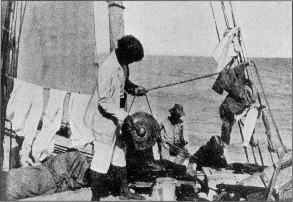 Black and white photograph of at least two men on a boat, with the horizon of the sky and sea in the background. A man is seen in the middle standing up and holding the handle of a circular gong in one hand, hitting its centre with a gong mallet in his other hand. He is wearing a sailor coat and knee-length light-coloured pants. Behind him is a man seated down wearing a fisherman’s hat which casts a dark shadow all over his head. He is wearing a long-sleeved top. On the left side of the image, sails can be seen, as well as clothes hanging onto a rope which stretches across the image to a line of ropes seen on the right which are tied in a grid-like pattern, stretched upwards beyond the frame. The grids are also attached with pieces of clothing or cloth tied on.