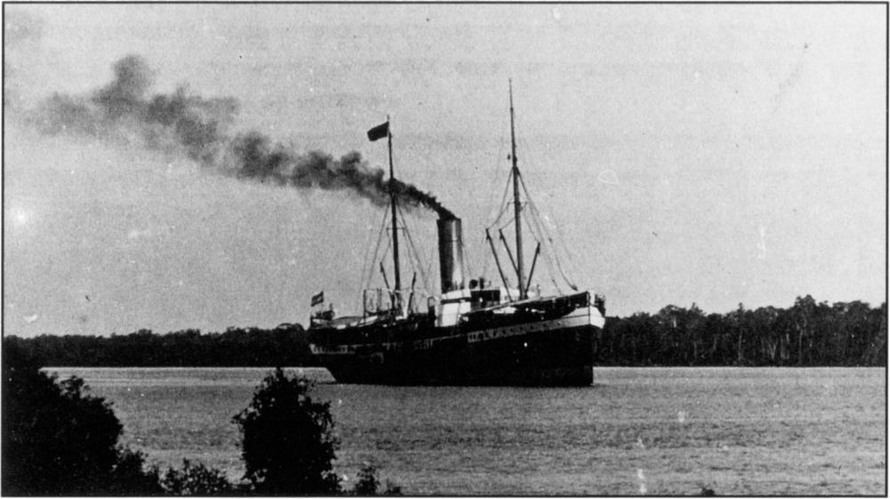 Black and white photograph of a large mail boat in the middle of the waters, with a dense line of trees in the background under the large plain sky. There is a dark-coloured flag raised to the top on a very high pole, and a trail of dark smoke coming out from its tall funnel or chimney. The mail boat appears to be sailing in the direction to the right of the image.