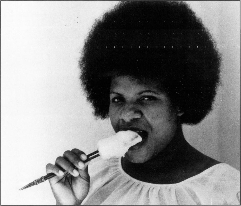 Black and white candid photograph of a dark-skinned Winimo village woman dressed in a white top with an impressively thick, tall and bushy head of hair, or afro, looking straight to the camera. With a stick of good amount of white stirred sago lumped to the head of heai or tongs in her hands and placed right at her mouth, she appears to be about to eat it. The heai is seen to have two very sharp and pointy ends which are bound together by twine. 