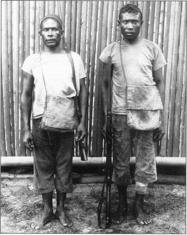 Black and white photography of two dark-skinned local bird hunters. Facing straight at the camera, the two young men are standing barefoot on dirt ground right in front a wall of tall bamboo branches which has a base of a larger horizontal bamboo branch as its base, they are both wearing plain light-coloured shirts and rolled up trousers with a rifle in hand and cross-body dirty fabric bag with thin handles and large open-flap as well as a headpiece or headband strapped on their heads. The man on the right appears to have a bandage wrapped over the bottom of his left calf. Their eyes are just slightly off the camera, with a bored and slightly concerned expression in their eyes.