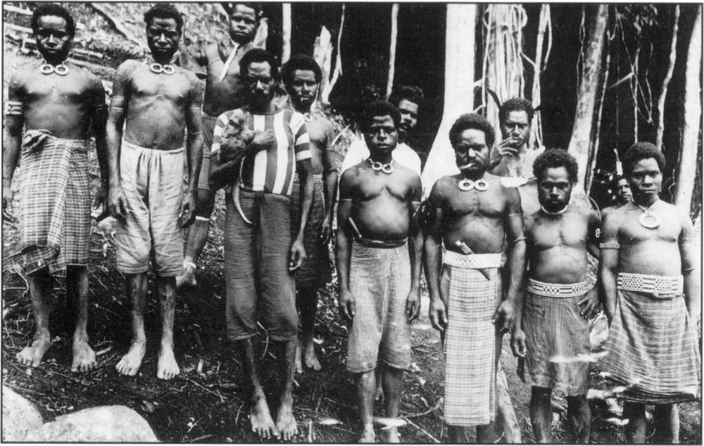 Black-and-white photograph of local bird hunters. They appear to have dark skin complexion, and are all bare-bodied with either a skirt, knee-length trousers or extended loin clothes covering their bottoms, except for one man who is standing off-centre to the right, who has on a shirt with thick vertical stripes. Most of them are donned in tribal necklaces and bracelets – many have on necklaces that have two large rings as pendants close resting right on their collarbones; one man to the farthest right has only one large ring pendant, with the man to his right having no pendants in sight. Barefooted and looking into the camera, the men appear to be standing right outside a hut with many trees on the side as seen in the background, with many little twigs sprayed on the dirt ground in which they stand on. Many men appear to have daggers stuck tied around their waists with a belt or the top of their skirt or trousers. Three of the men have cigarettes in their mouths and are appearing to be smoking. The man in the striped shirt appears to be holding an animal in his arm. 