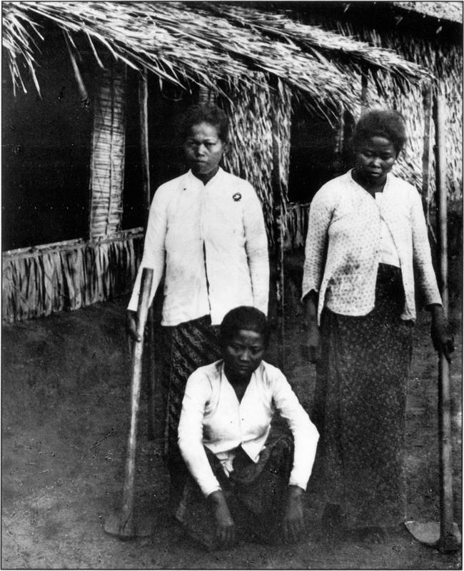 Black and white photo of three female Indonesian tobacco plantation employees, two of whom are standing in front of a straw house also made with leaves with a long flat-blade shovels in their hands while the other is squatted with her arms resting on her propped knees. They are all wearing long-sleeved cardigan or tops, with at least the top two buttons buttoned up, and are all in long maxi printed skirts. They appear to have very short, boyish and thick hair. One of the women standing to the left is looking straight into the camera with a slight frown, while the other two have their eyes peered onto the ground. None of them look very happy – almost as if despondent. 