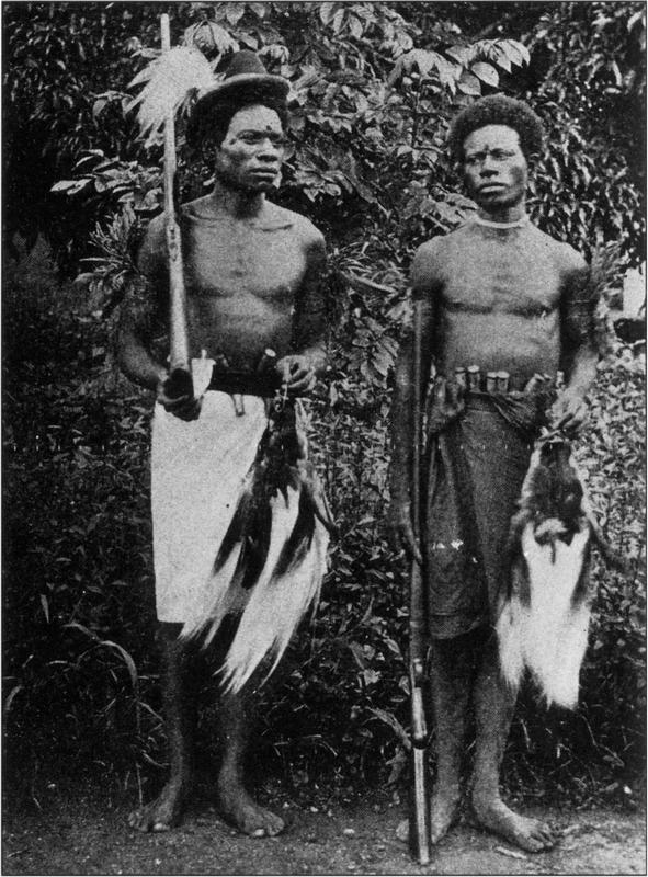 Black and white photograph of two local, bare-bodied and bare-footed birds of paradise hunters with rifles in each of their hands standing on the dirt ground in front of a mass of leaves or bush. They are both donned in knee-length wrap skirts, with the man on the left wearing a light-coloured one and the other dark, with a belt holding onto several short, thick wooden tools tucked in. The man standing to the left of the frame has a small and thick fedora-shaped hat on, with a bunch of light-coloured plumes attached on its side. He is also holding the foot of a tall rifle in his right palm as its forearm rests on his shoulder, with the barrel perched sky-ward. On his other hand, he clasps a dead Lesser Bird of Paradise which is hung by a small hooped string on its head, with its long dark body and light-coloured plumes suspended lifelessly. The man has two distinct dark round marks on his forehead right above his nose bridge, and one to the side of his right eye by his temple. He is facing to the right direction, looking away from the camera with a rather sombre look on his face. The man to the right is looking to the other side towards the left. He has short, thick afro hair, and also has a distinct round mark on his forehead. He dons a piece of necklace tied closely to his neck. On his right stands a rifle which is supported closely by his hand. On the other hand is also a dead Lesser Bird of Paradise hanging lifeless as a soft white bunch of plumes.