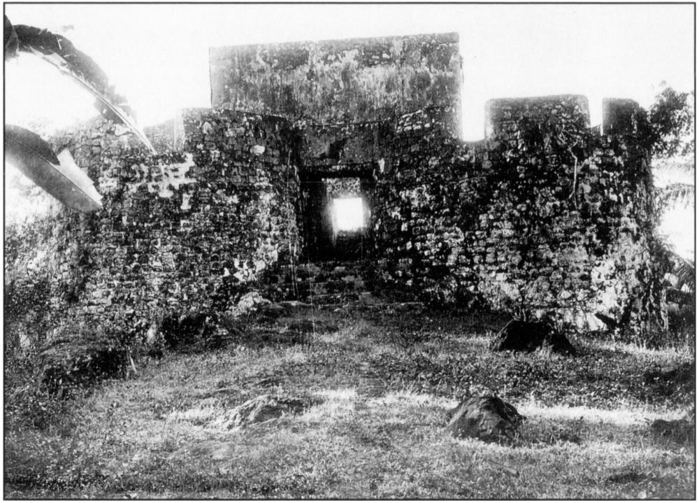 Black and white photo with the sky brightly exposed and constituting the top-fifth portion of the image, serving as backdrop to the ruins of a portion of an old bricked Portuguese fort. The fort stands on a grassy area pimpled by a few ground rocks which foregrounds the image. The top of fort is seen to have parapeted outlines with low-walled battlements. Right in the middle of the depicted portion of the fort marks a squared-out opening through a doorway elevated by a few steps, revealing a peephole of the bright background sky. Some palm leaves creep into the shot on both sides, indicating the presence of trees around the fort. 