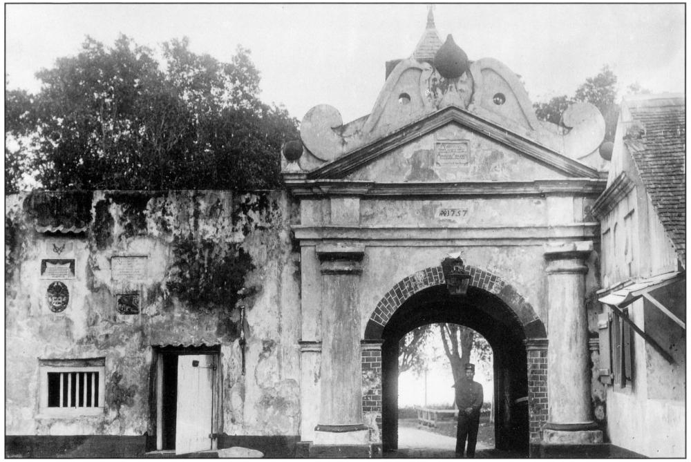 Black and white photo of a Dutch fort gate with stone bulwarks and thick walls. The walls appear to have signs of age, with dark patches of varying shades as well as several plaques with inscriptions spaced around on the front-facing walls. Towards the bottom left corner of the fort gate is a rectangular barred window, with an outer-left bar apparently missing, thus leaving a notable gap next to the five evenly-spaced vertical bars. Next to the barred window is a normal-sized door which is left ajar. Behind this section of the wall is the top-half of a large tree. The arched roof section of the tall main entrance is adjoined to the right, with a full rainbow-shaped, curved edge of brick patterns hovering over a uniformed guard standing slightly off-centre to the right. He is wearing a flat-top hat, long-sleeved shirt buttoned to the top with two breast pockets and a pair of trousers. He appears to have a darker skin complexion and a moustache. With his hands crossed in front of his body, he appears to have a very austere semblance. Behind him is a sneak peek to what lies behind the fort gate: a significant portion of a fenced tree trunk, with some leaves sprawling over against the bright sky. Above the brick-patterned arch marks the numbers “1757”, indicating the year in which the fort gate was either constructed or established. Above that is a triangular top, capped by a graphical moustache-like shape with an inverted top-like shape perched in the middle, giving the fort gate its distinctive appearance. A steeple-like structure peers behind slightly in the background. Adjacent to the arched entranceway is a portion of a building of similar height and pointed bricked roof, with an opened planked window on the first level and three indented shapes on the second level. 