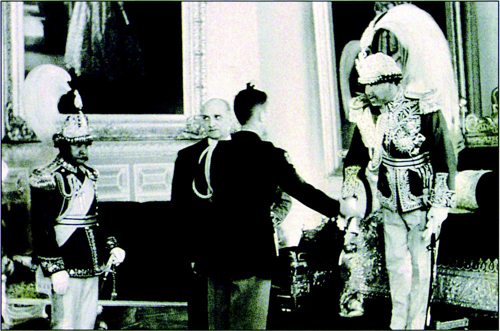 Black and white photograph showing the Prime Minister of Nepal standing on the left side of the picture with an embroidered uniform top and royal crown adorned with a huge interwoven flank plumes. The King, on the other right side of the frame, is also donned a royal crown with larger plumes and a more embroidered and embellished uniform top. The King is seen shaking hands with the American Ambassador, who stands on the centre of the image, with his back to the camera. The American Ambassador is wearing a black uniform with a waist belt and lighter-coloured trousers, and braided sash on his left arm. In front of the American Ambassador is another man eyeing towards the top-left of the image. In the room there are very large portraits hung on the walls with ornamental frames. There also appears to be regal furniture such as sofas adorned with a sort of embroidery and silver, as well as what appears to be the King’s throne on the right end of the picture, positioned right behind the standing and engaged King.