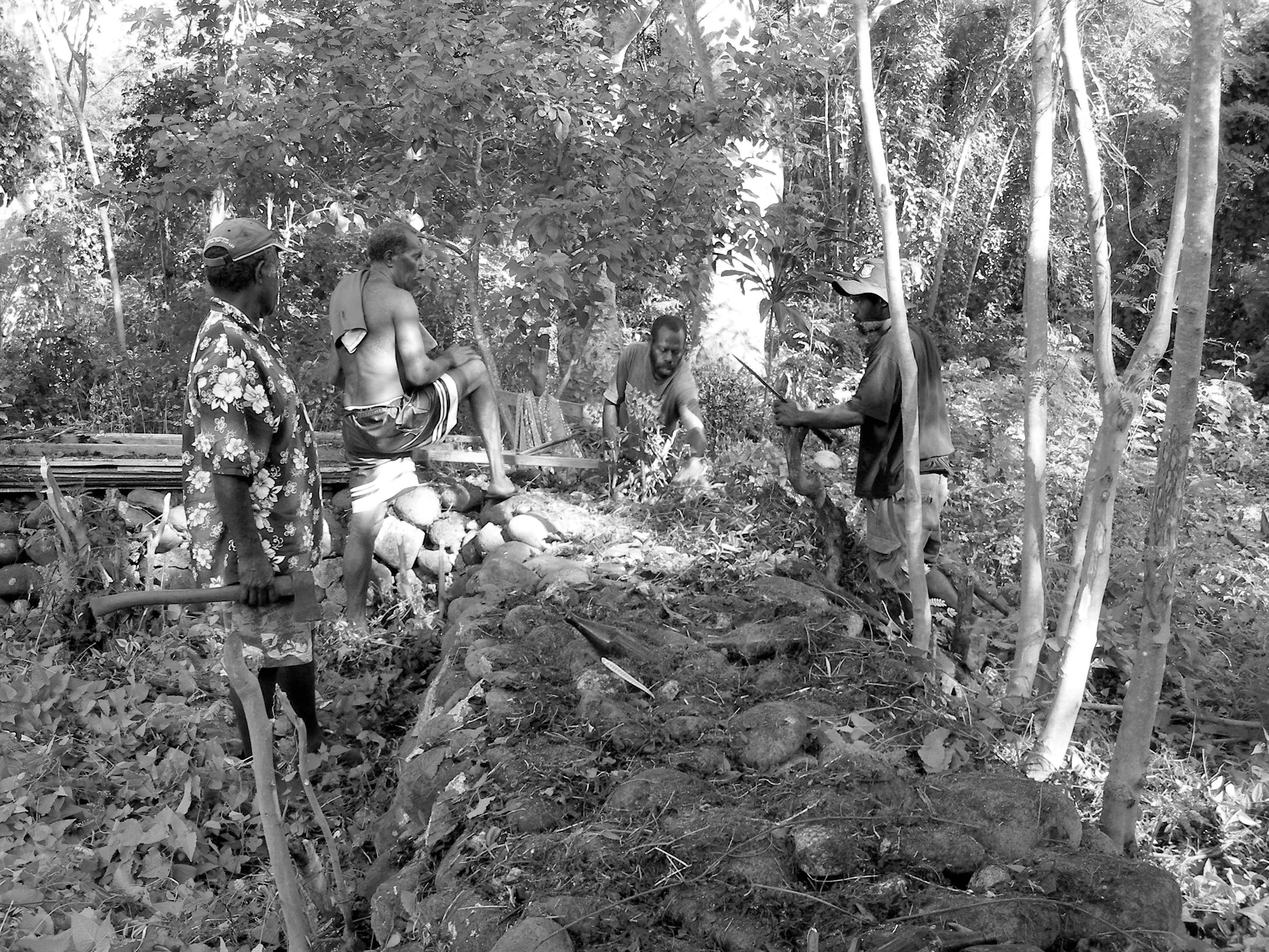 Black and white image of four men working on an archaeological site surrounded by thick bush.