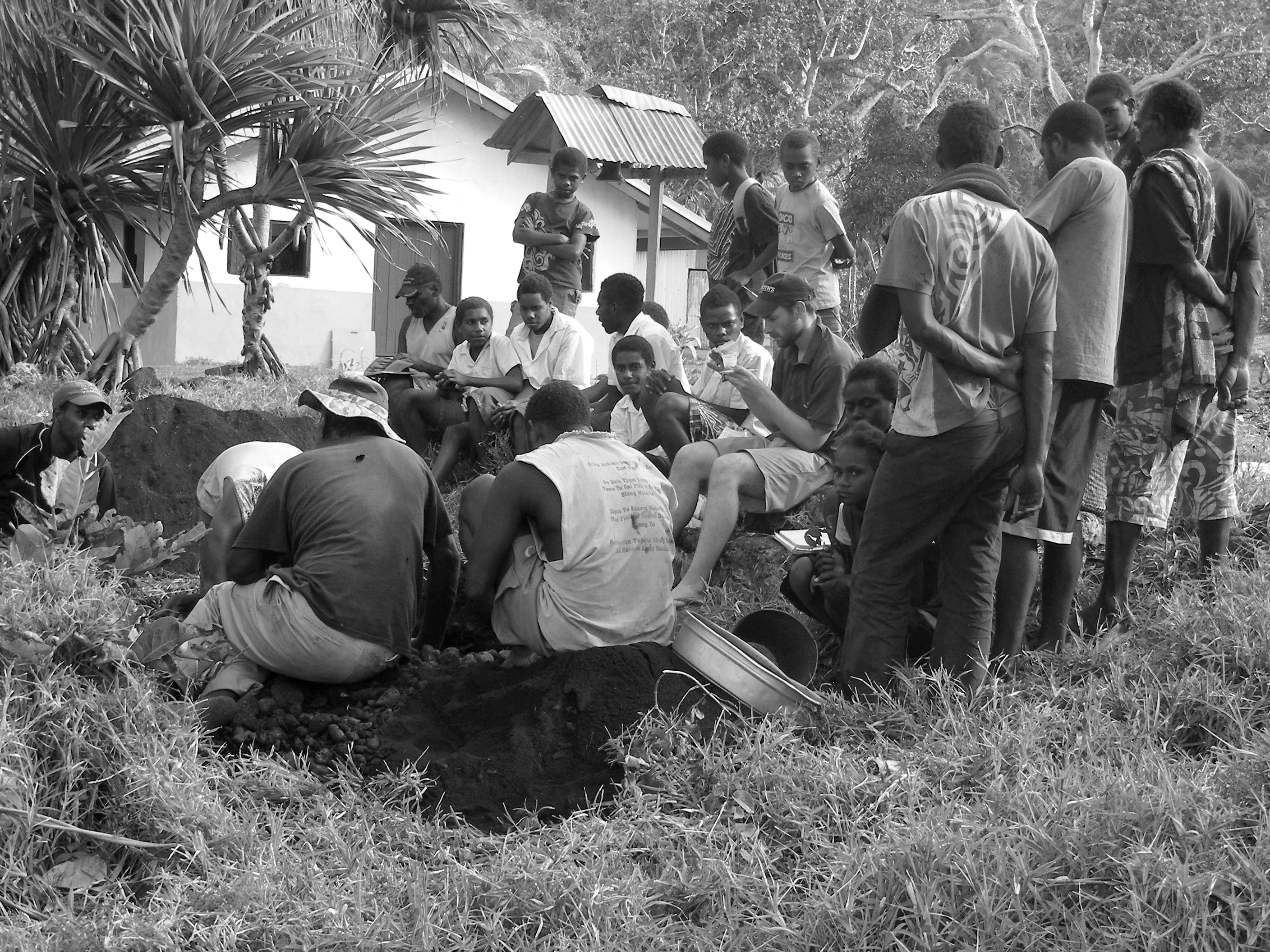 A large group of people gather around two men working on an archaeological dig. A white house is visible in the background.