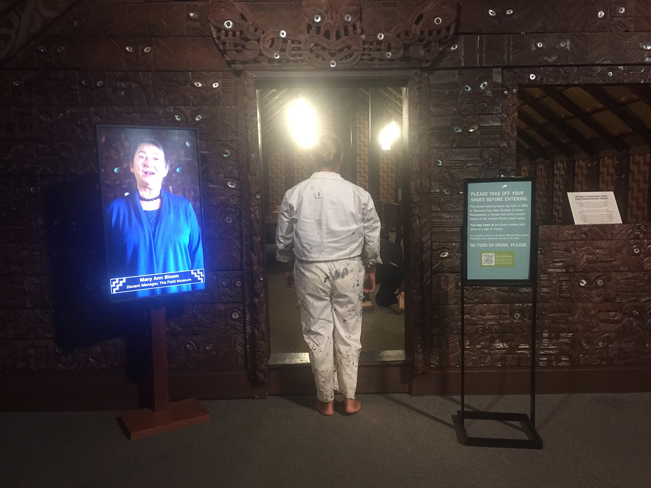 With his back to the camera, Jack Gray, wearing his deceased father's painting overalls, stands in front of a doorway to a building (Ruatepupuke II), about to enter. There is museum signage to his right including one placard on a stand. On his left there is a portrait of a woman.
