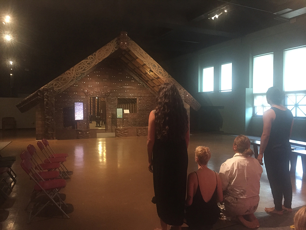 Two dancers sit and two stand in the foreground with their backs to the camera, poised to begin a welcome dance for manuhiri (visitors). There is open floor in front of them and a house at the other end of the room within the Field Museum. There are empty seats and benches lined up on either side of the dancers.