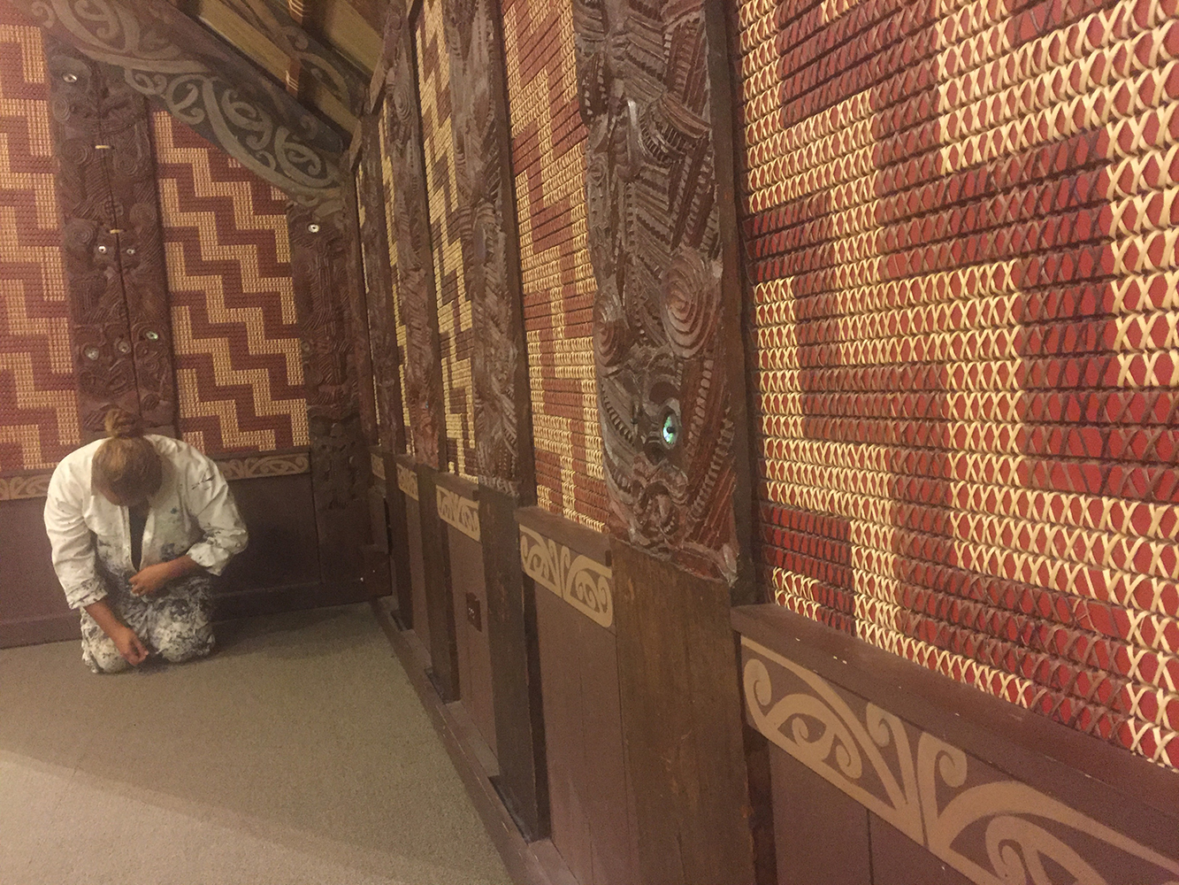 Jack Gray kneels in a corner of the house, Ruatepupuke II, head down and back against one wall. Decorative woven fabrics and carvings cover these interior walls.