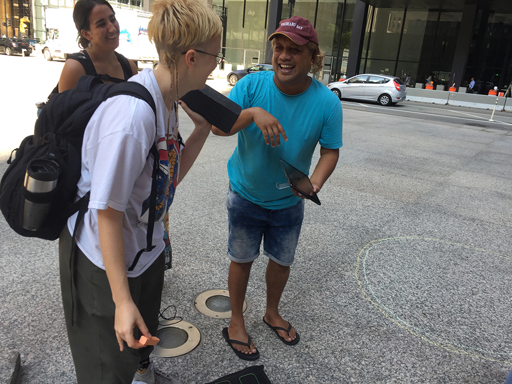 Three people laugh and smile in the foreground against an urban Chicago streetscape. The woman closest wears a backpack with a thermos visible in the side pocket. Concrete stretches into the background where there is a road with cars on it.