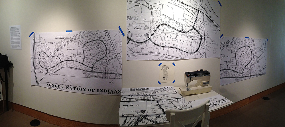 An installation of fabric and paper from Seneca relocation maps in a museum. The materials are plastered to a white wall and there is a table with a section of map on it and a sewing machine.