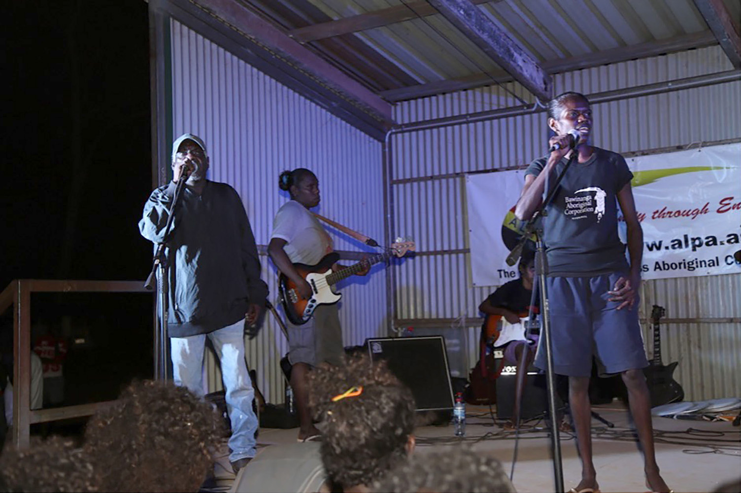 A band performs on a stage with tin walls. David Maxwell (left) and his daughter, Stephanie Maxwell James (right) sing into microphones. Behind them is Rona Lawrence on bass guitar and Tara Rostron on electric guitar.