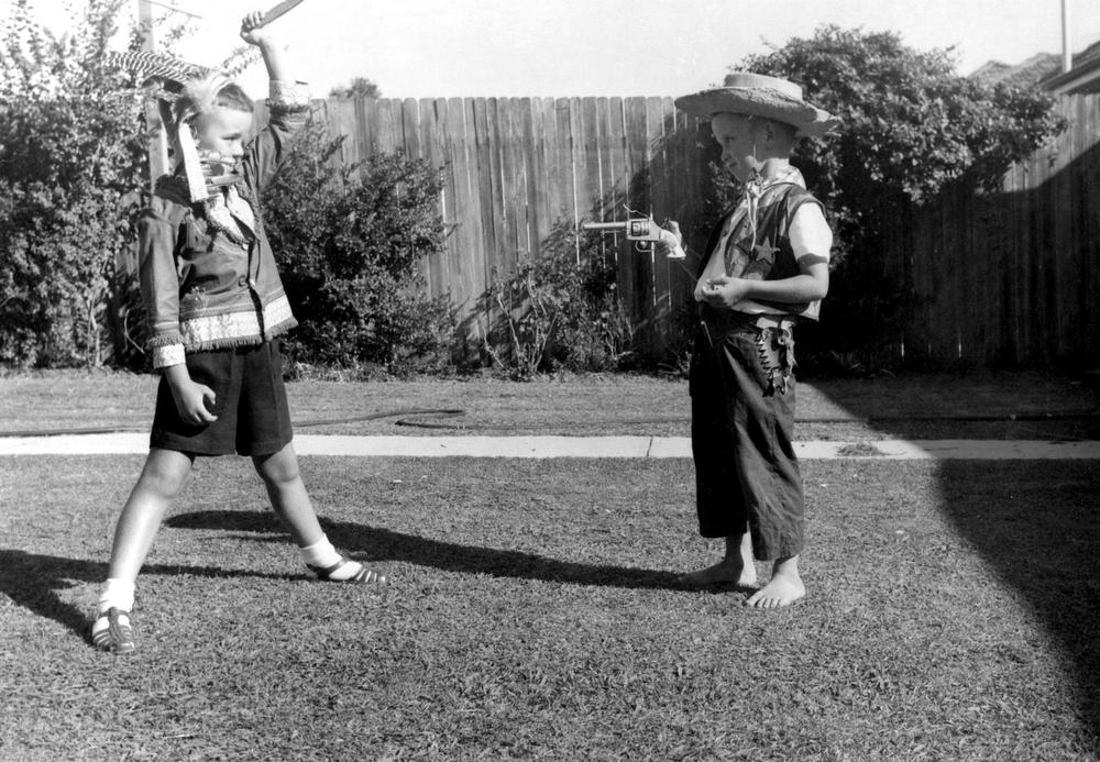 Black and white photo of two little boys, between the ages of five and seven, standing in profile, facing one another in a backyard. The boy on the left is wearing a feathered headdress, a fringed button-up shirt, dark shorts and socks with sandals. He raises a toy weapon high above his head towards the little boy on the right, who is wearing a cowboy hat and a vest with a sherriff's badge, along with dark trousers and no shoes. The boy on the right points a gun at the boy on the left, and carries a walkie talkie in his pocket. Both boys are smirking.