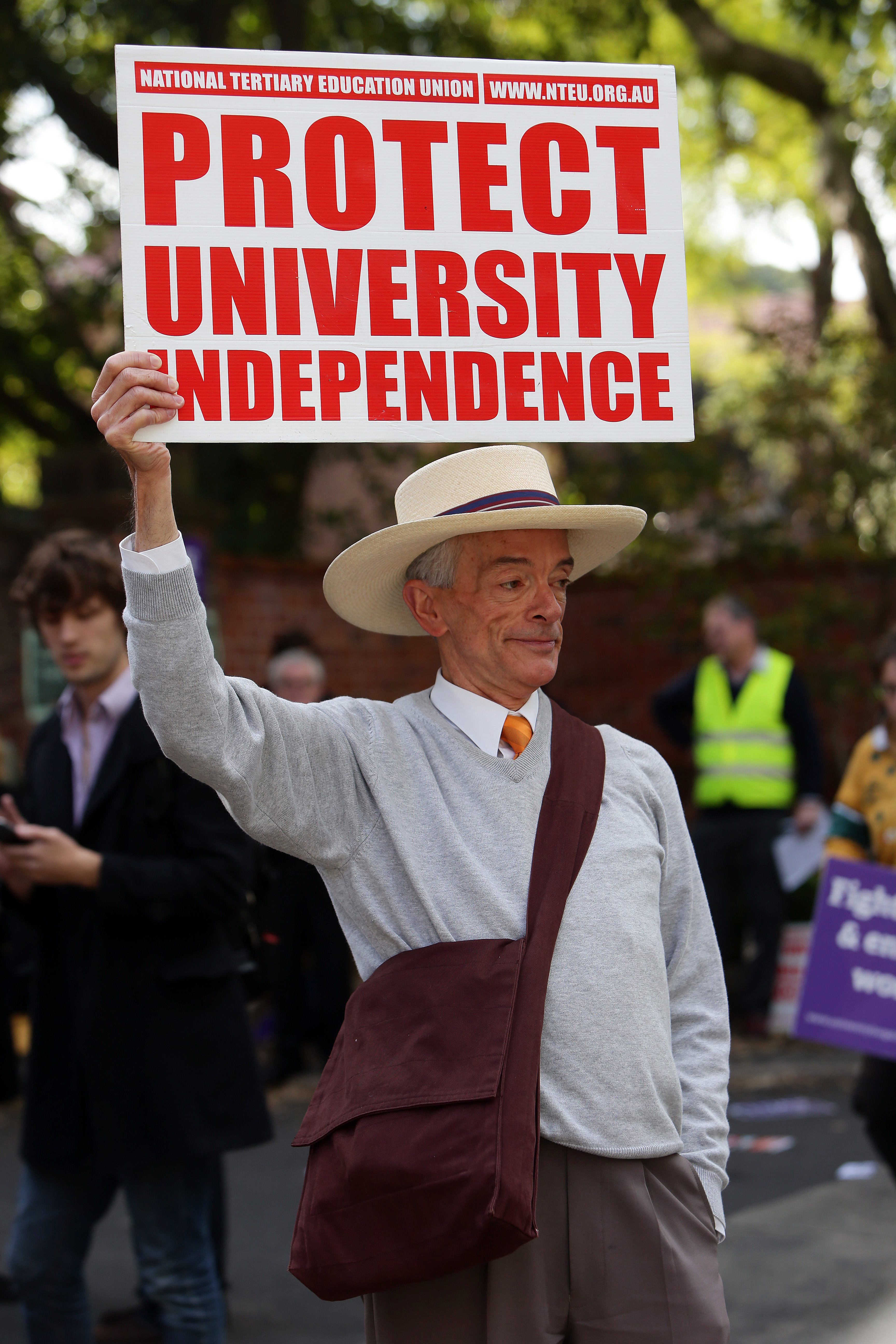 Black and white photo of a man in a boater hat, wearing a sweater over a white collared shirt and tie, and a satchel bag slung across his body from his left shoulder to the opposite hip. In his right hand, he holds up a large sign that reads "Protect University Independence" with the web address for the National Tertiary Education Union. His left hand is tucked in the pocket of his slacks. The man has a serious expression on his face and looks off-camera to his left. There are several other people blurred in the unfocused background.