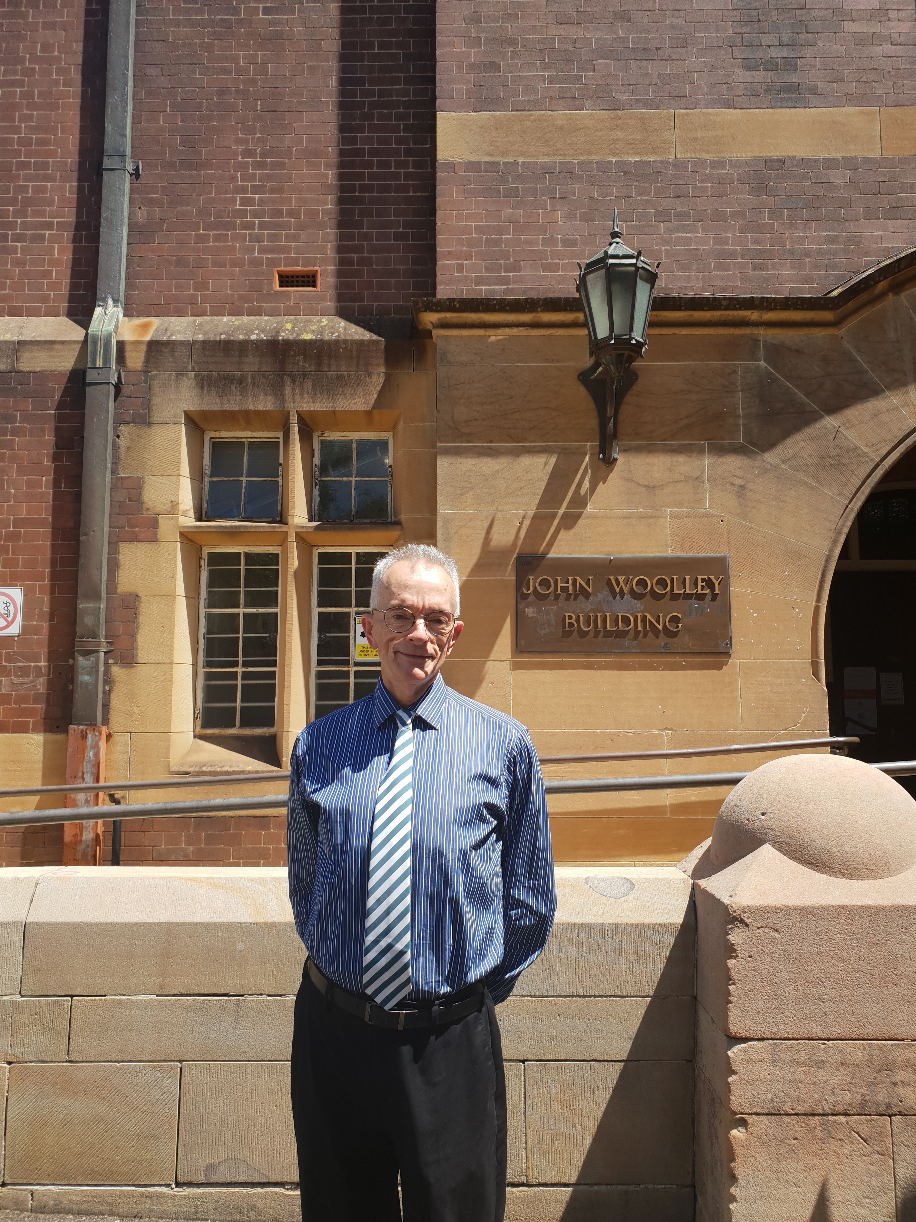 Black and white photo of a man with short grey hair, smiling and standing with his hands tucked behind his back, in front of a stone wall with a plaque reading "John Woolley Building". The man is wearing glasses, along with a pin-striped long-sleeved button-up shirt, a diagonally striped tie and dark slacks. 