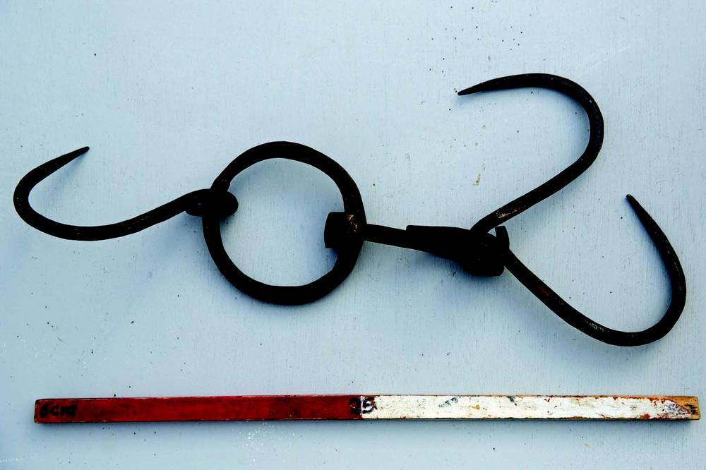 An old, rusted iron tool with two large curved hooks connected by a central ring and short chain links, placed on a light grey surface. Below the tool is a red and white measuring stick marked with "0 cm" on the left side.