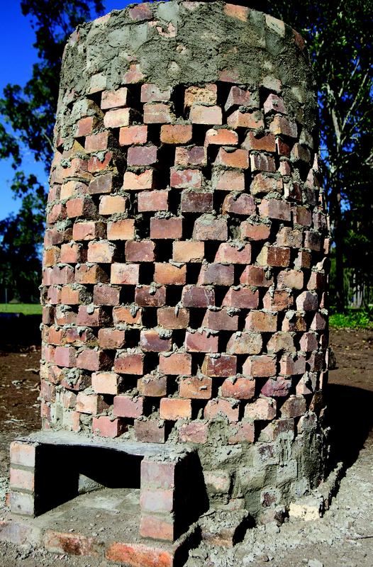 A cylindrical brick structure with a rough, unfinished top and an open rectangular cavity at the base. The bricks are arranged in a staggered pattern with gaps between them, creating a ventilated design. The structure sits on bare earth with trees and blue sky in the background.