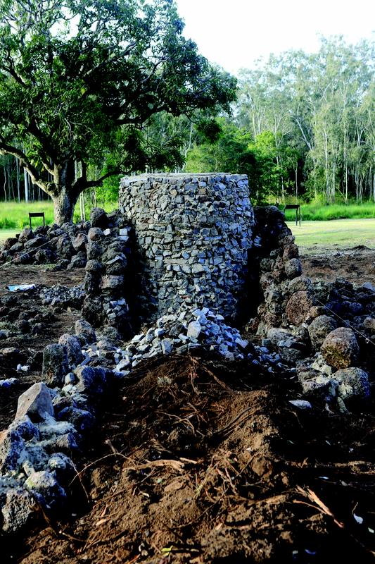 A cylindrical structure of stacked stones, with a rough, uneven surface. It has two side buttresses and a pile of loose stones leading away from its base. The structure sits on bare earth in an open area with scattered rocks, with a large tree and grassy field in the background.