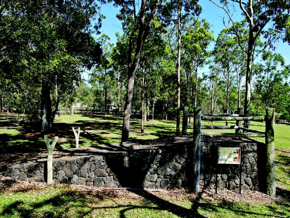 A low stone wall made of dark volcanic rocks, bordered by upright timber posts and horizontal rails forming a simple fence. There is an information sign mounted on two metal posts in front of the wall. The setting is a grassy park with tall eucalyptus trees and dappled sunlight.