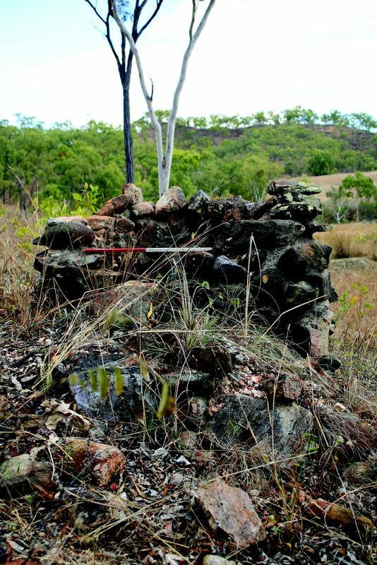 Colour photograph of a partially collapsed stone structure in a grassy landscape with scattered trees. The stones are dark and irregular, stacked to form a low wall. A measuring rod lies horizontally across the structure. The background shows hills and sparse vegetation under a cloudy sky.