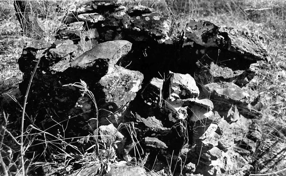 Black-and-white photograph of a collapsed stone structure made of irregular rocks stacked loosely. The structure is surrounded by dry grass and vegetation, suggesting an outdoor rural setting. The stones show weathering and rough textures.