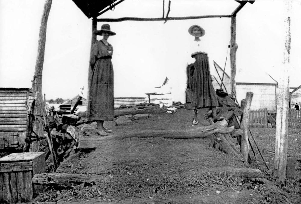 Historic black-and-white photograph showing two people standing on a raised earthen platform under a simple timber frame structure. The platform is surrounded by rough wooden fencing and farm buildings in the background. The individuals wear long garments and wide-brimmed hats, and the setting suggests an early rural or agricultural environment.