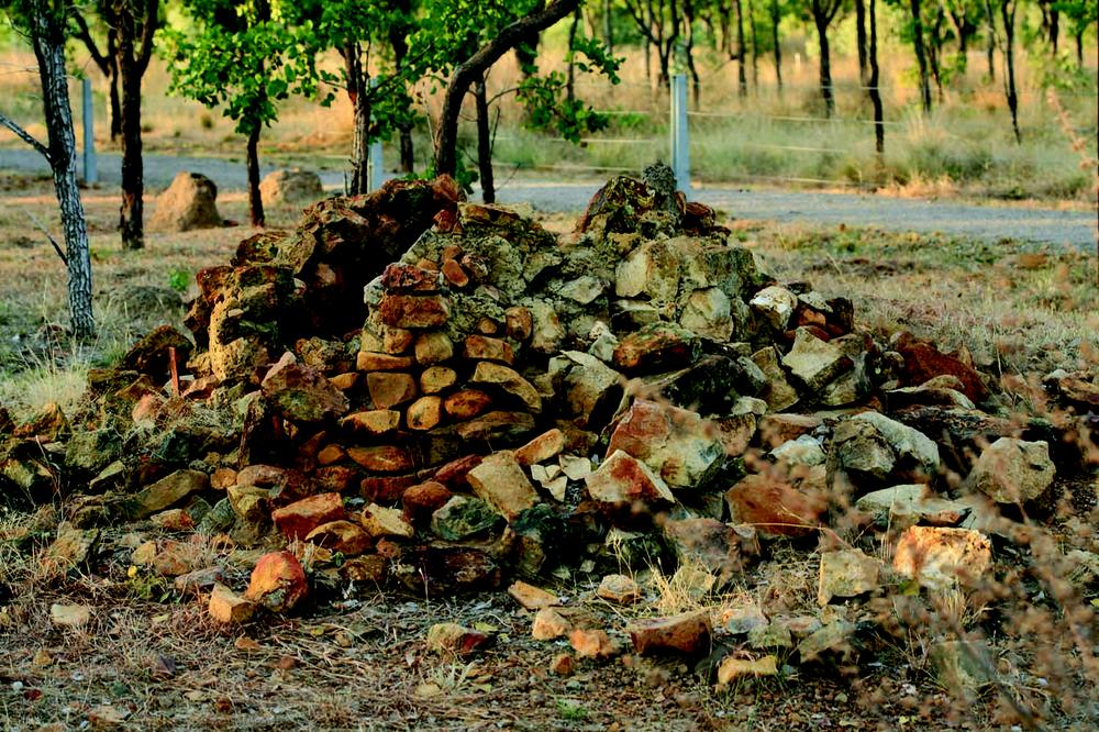 A collapsed stone structure made of irregular rocks stacked loosely, forming a mound. The structure sits in a dry, grassy area with scattered trees and a dirt track visible in the background. The stones are weathered and vary in size and colour.