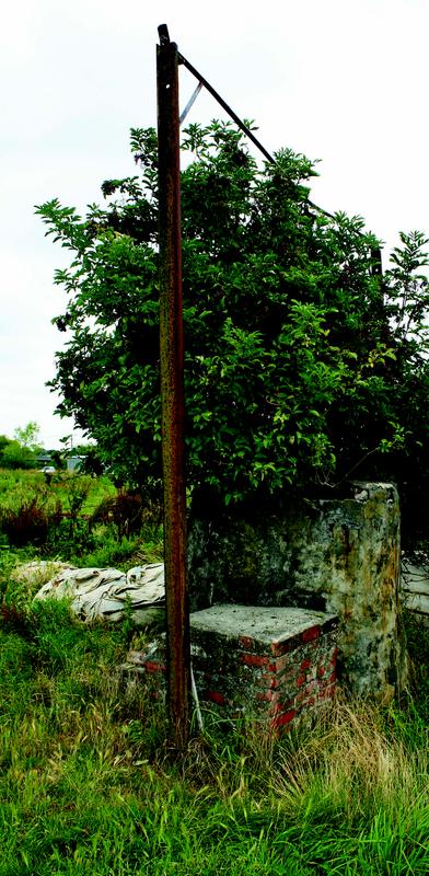 A weathered well structure in a grassy field. The well consists of a square stone and brick base with a rusted metal frame extending vertically and curving at the top, possibly for a pulley. Dense foliage grows behind the structure, partially obscuring it. The background shows open grassland and scattered vegetation under a cloudy sky.
