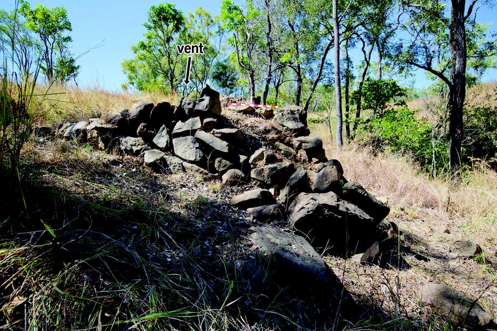 A partially collapsed stone structure built from irregular rocks stacked loosely on a sloping hillside. A super-imposed arrow points to the top left side of the structure with the word "vent" above it. The surrounding area is dry with tall grass and scattered trees under a clear blue sky.