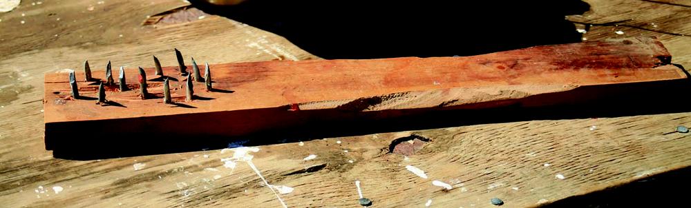 Close-up of a handmade wooden tool resting on a weathered timber surface. The tool is a rectangular block of wood with two rows of bent metal nails protruding upward, likely used for scraping or combing. The wood shows signs of wear and rough carving, and the table beneath has paint splatters and nail heads visible.