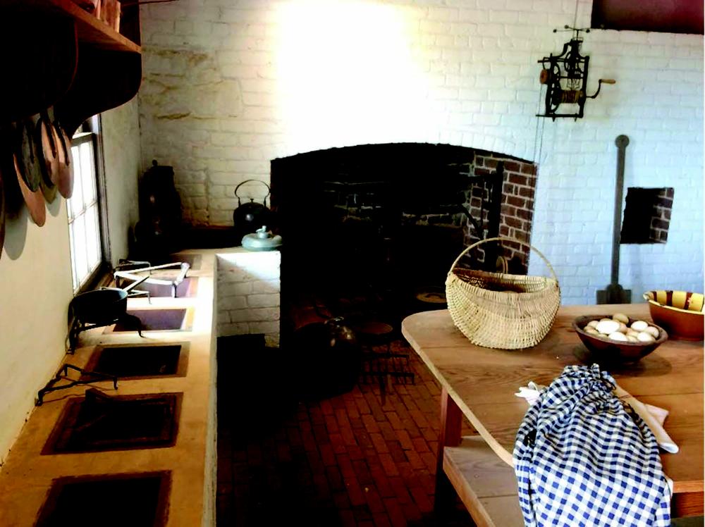 A historic kitchen with a large brick fireplace and whitewashed walls. The hearth contains iron pots and kettles, with additional cookware on a side bench featuring built-in openings for cooking. A wooden table in the foreground holds a wicker basket, a bowl of eggs, and a checked cloth. Copper pans hang on a shelf near a window.