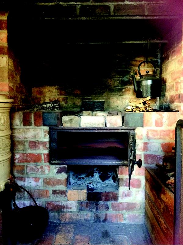 A brick fireplace with a built-in iron oven and soot-stained bricks. Two metal kettles hang from a horizontal rod above the firebox, and pieces of firewood are stacked on either side. A black cast iron pot sits on the floor, and a wooden box filled with logs is visible to the right.