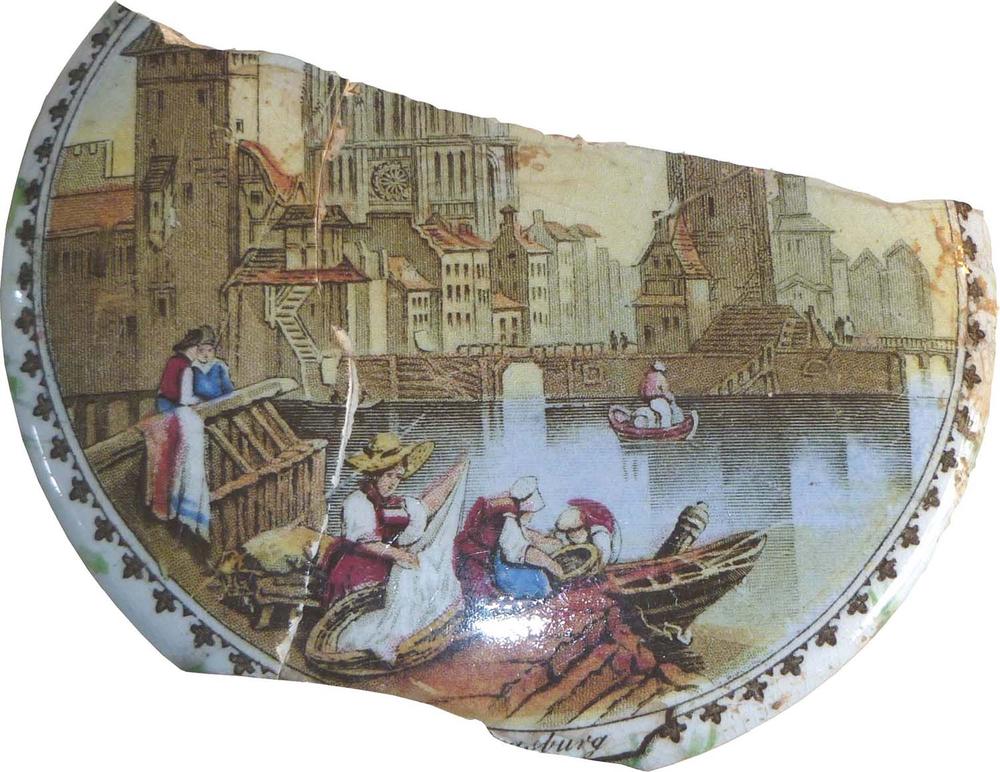 A broken piece of decorative ceramic featuring a detailed coloured illustration of a riverside scene. The design shows people in period clothing working near wooden boats on the water, with tall historic buildings and a cathedral in the background. The fragment has a curved edge with a floral border pattern.