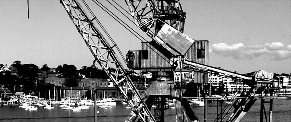 A black and white photograph of a crane in the forefront and the harbour in the background.