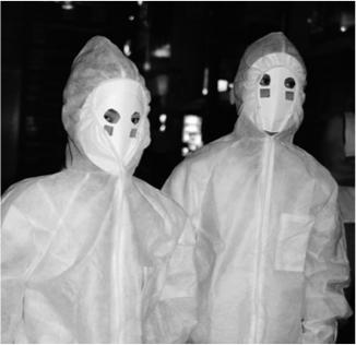 A black and white photograph of two people in white medical suits and white masks. 