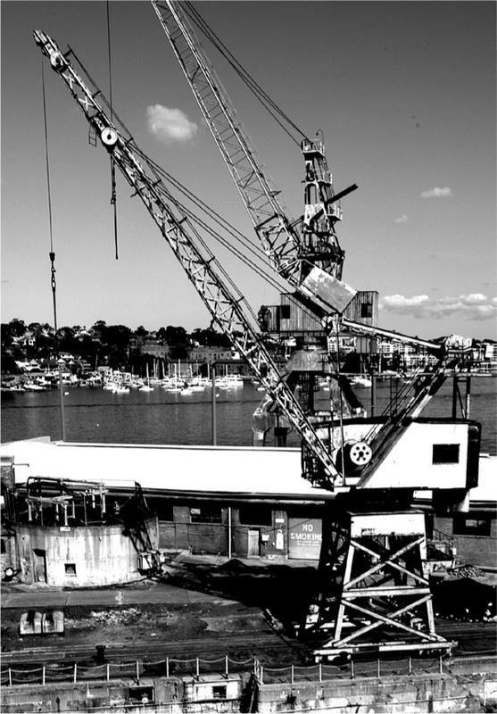 A black and white photograph of a crane being used in construction. There is the harbour and ships in the background.