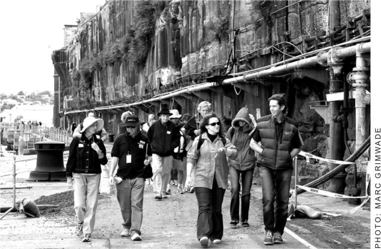 A black and white photograph of a group of men and women walking around Cockatoo Island and listening to an historical tour.